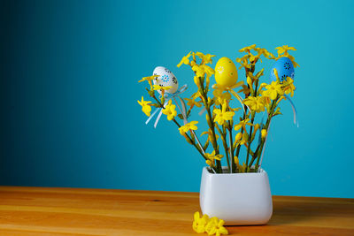 A bouquet of spring yellow flowers on blue background with easter eggs with pattern in a white vase.