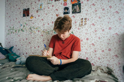 A teenage girl sitting on a bed draws in a sketchbook.