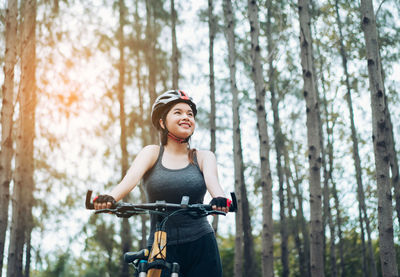 Low angle view of happy female cyclist standing with bicycle in forest