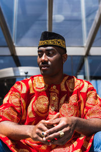 Serious adult african guy in traditional red clothes and kufi cap looking away while sitting in city street near building with glass walls in sunny day
