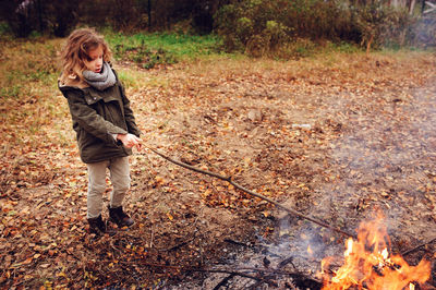 Full length of girl holding stick on bonfire while standing in forest during autumn