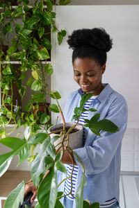 Young african american woman with wide smile holds house plant and examines philodendron flower