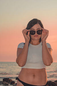 Portrait of woman wearing sunglasses while standing at beach against sky during sunset