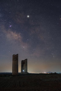 Anonymous person with lantern light on ancient stone towers on empty sandy ground under dark starry sky with milky way