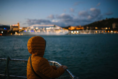 Rear view of man looking at sea against illuminated city