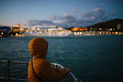 Rear view of boy looking at sea against illuminated city