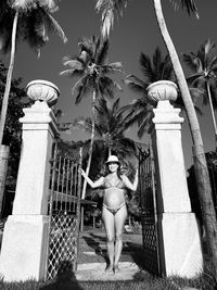 Pregnant woman in bikini standing on gate against by palm trees