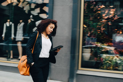 Mid adult businesswoman walking while using digital tablet with hand in pocket against building in city