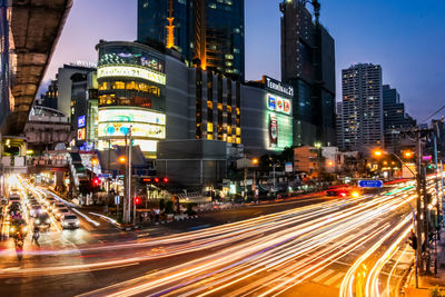 Light trails on city street by buildings against sky at night