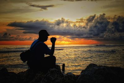 Silhouette man gesturing while sitting on rock at beach during sunset