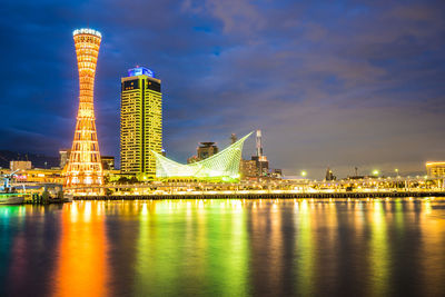 Illuminated modern buildings by river against sky at night