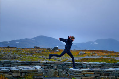 A young woman leaps over a gap in a low stone wall at a rest stop on sognefjellet .