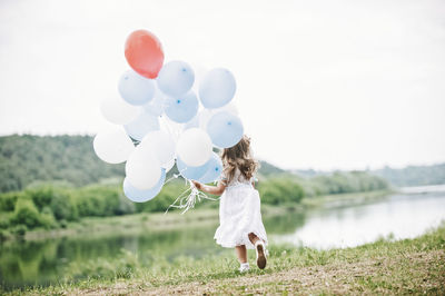 Rear view of girl holding balloons while walking at lakeshore against sky