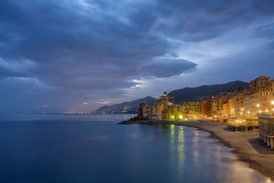 Illuminated buildings by sea against cloudy sky