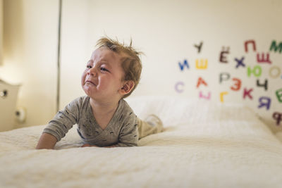 Crying baby on bed with white blanket and alphabet on wall behind him