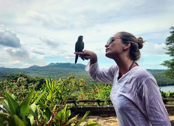 Woman looking at parrot on finger against sky