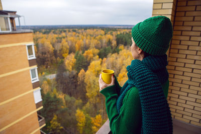 Woman in a green knitted sweater and a hat stands with a yellow mug on the balcony, autumn forest