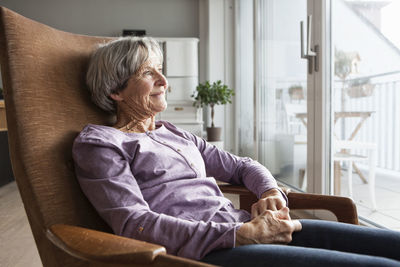 Portrait of senior woman sitting on armchair at home looking through window