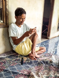 Full length of man using smart phone while sitting against wall at home