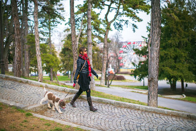 Woman walking with dog on footpath by trees during winter