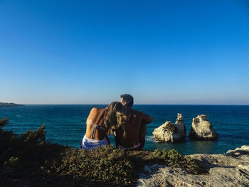 Friends sitting on rocks by sea against clear blue sky