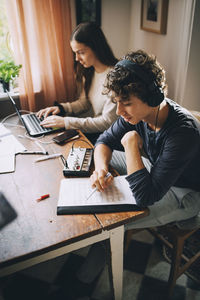 High angle view of teenage boy listening music with headphones while friend using laptop at table in house