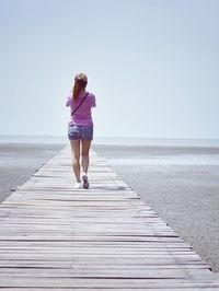 Rear view of woman walking on boardwalk on beach against clear blue sky