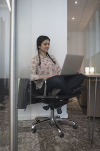 Young woman sittin cross-legged in office typing on her laptop.