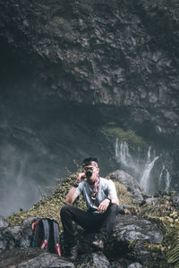 Young man sitting on rock against waterfall