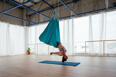 Side view of active female practicing swan dive in hammock during aerial yoga training in light spacious studio with mat