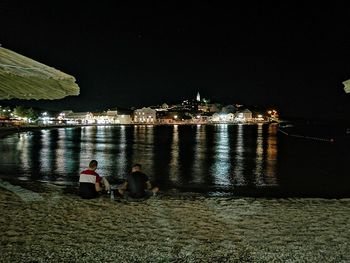 People sitting by river against illuminated city at night