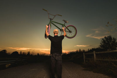 Rear view of man lifting bicycle against sky during sunset