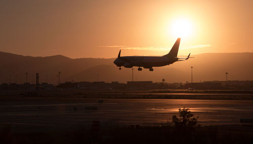 Silhouette airplane flying over runway against sky during sunset