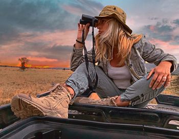 Woman looking through binoculars while sitting on car roof against sky during sunset
