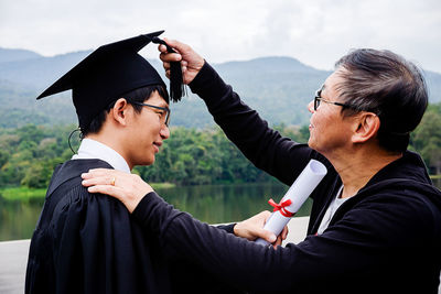 Father adjusting mortarboard of son against mountain