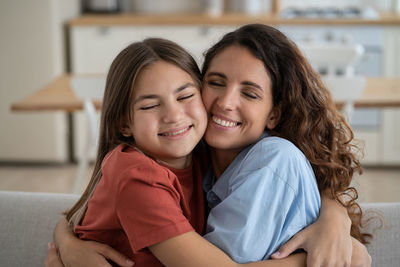 Close-up smiling positive mother and daughter hugging each other tightly sits on sofa in apartment