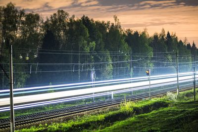 Light trails on road by trees against sky during sunset