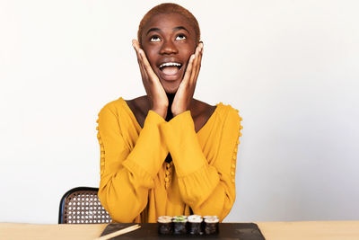 Young woman with sushi on table looking up against white wall at home