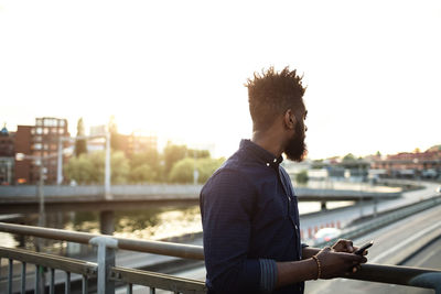 Young man with mobile phone standing on bridge against sky