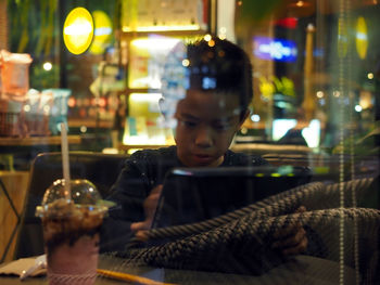 Boy using digital tablet in illuminated cafe seen through window at night