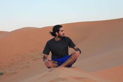 Young man sitting on sand dune in desert against clear sky