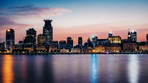 Illuminated buildings by river against sky during sunset