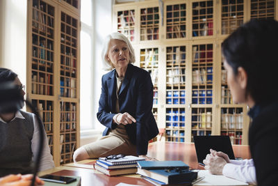 Senior professional gesturing while sitting on table during meeting in library