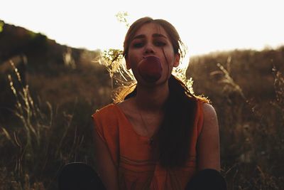 Portrait of young woman eating apple while sitting on field against clear sky during sunset