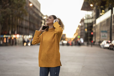 Happy woman holding mobile phone enjoying music through wireless headphones on footpath