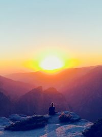 Rear view of woman sitting on cliff by mountains against sky during sunrise