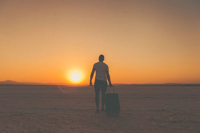 Rear view of man walking with suitcase on snow field against sky during sunset