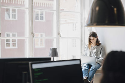 Focused young female programmer using laptop while sitting by window in creative office