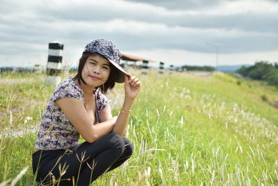 Portrait of young woman wearing cap crouching on grassy field against cloudy sky