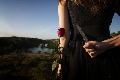 Midsection rear view of woman holding knife and rose behind her back on land